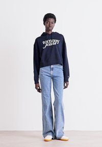 Persona che indossa un hoodie navy di Tommy Jeans, jeans a campana di un blu chiaro e sneakers bianche con suole in gomma, in piedi davanti a uno sfondo bianco semplice.