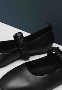 Zwarte leren ballerina's met een gladde afwerking en een ronde neus, voorzien van een gestructureerde, gekreukte band over de bovenkant.