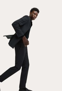 Czarny garnitur o dopasowanym kroju, w połączeniu z czarną koszulą. Model idzie z lekkim pochylonym ciałem, prezentując gładką teksturę i wyrazisty design garnituru.