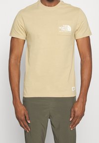 Muž v béžovém tričku s krátkým rukávem The North Face s logem na kapse na hrudi a olivově zelených kalhotách.