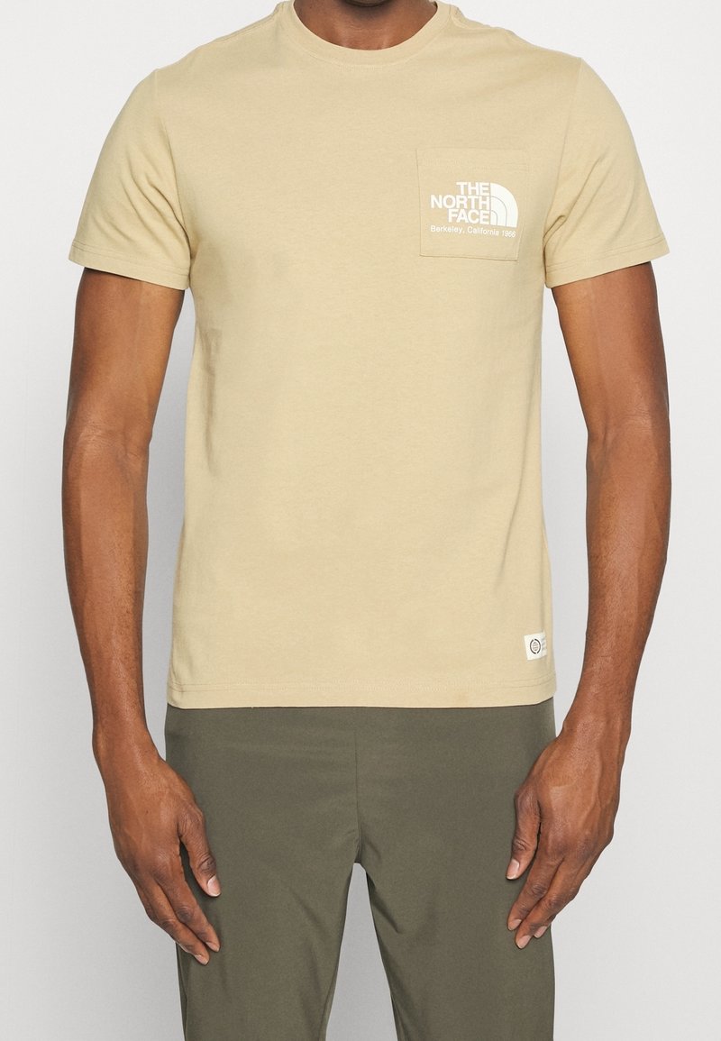Muž v béžovém tričku s krátkým rukávem The North Face s logem na kapse na hrudi a olivově zelených kalhotách.