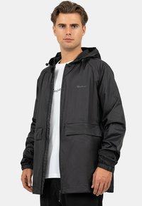 Zwarte lichtgewicht jas met capuchon, een ritssluiting aan de voorkant, elastische manchetten en twee steekzakken. Voorzien van subtiele branding op de borst.