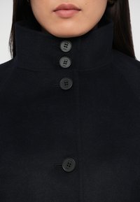 Tumši jūras zila vilnas mētelis ar augstu apkakli, pieciem melniem pogām un gludu tekstūru. Pogām ir iegravēts "moje PARIS".