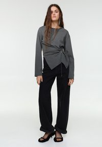 Siv dolg rokav s topom z asimetričnim sprednjim dizajnom in stranskim vrvico, kombiniran s črnimi hlačami širokih hlačnic in črnimi mehki sandali.