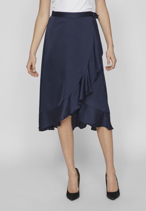 Jupe en satin bleu marine avec des volants superposés à l'ourlet et un détail de lien sur le côté. Texture lisse, design jusqu'aux genoux, associée à des talons noirs.