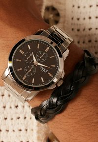 Silver metal watch with a black dial, three chronograph subdials, and silver markers. Worn alongside a braided black leather bracelet.