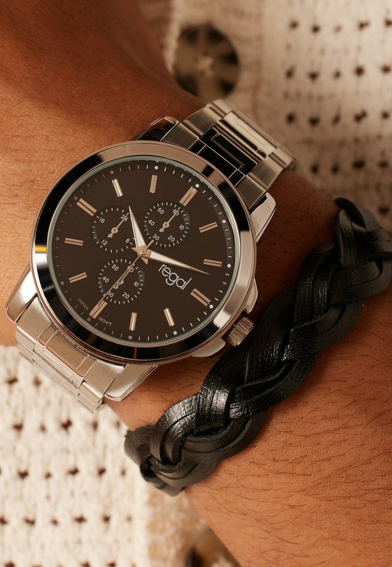 Silver metal watch with a black dial, three chronograph subdials, and silver markers. Worn alongside a braided black leather bracelet.