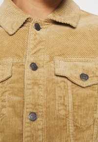 Giubbotto in velluto a coste di un leggero color beige, con un motivo a coste testurizzato, bottoni metallici, colletto e due tasche frontali con chiusura a pattina.