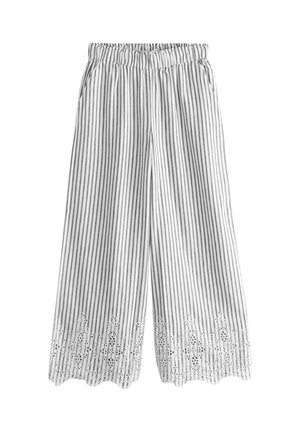 Femeie stând desculță pe o plajă cu nisip lângă stânci, purtând un top fără mâneci în dungi alb și albastru și pantaloni largi cu detalii perforate.