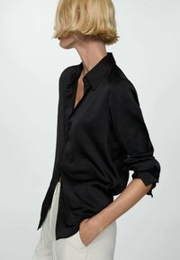 Blouse en satin noir avec un col classique, un devant à boutonnière et des manches retroussées, associé à un pantalon couleur crème. Texture lisse, design élégant.