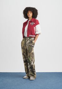 Rode en witte varsityjas met embleem, gecombineerd met wijde camouflagebroek. Model draagt grijze sneakers. Effen achtergrond.