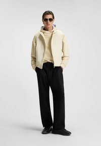 Giacca zip con crema sopra un hoodie beige chiaro, abbinata a pantaloni larghi neri e sneakers nere. Gli accessori includono occhiali da sole scuri.