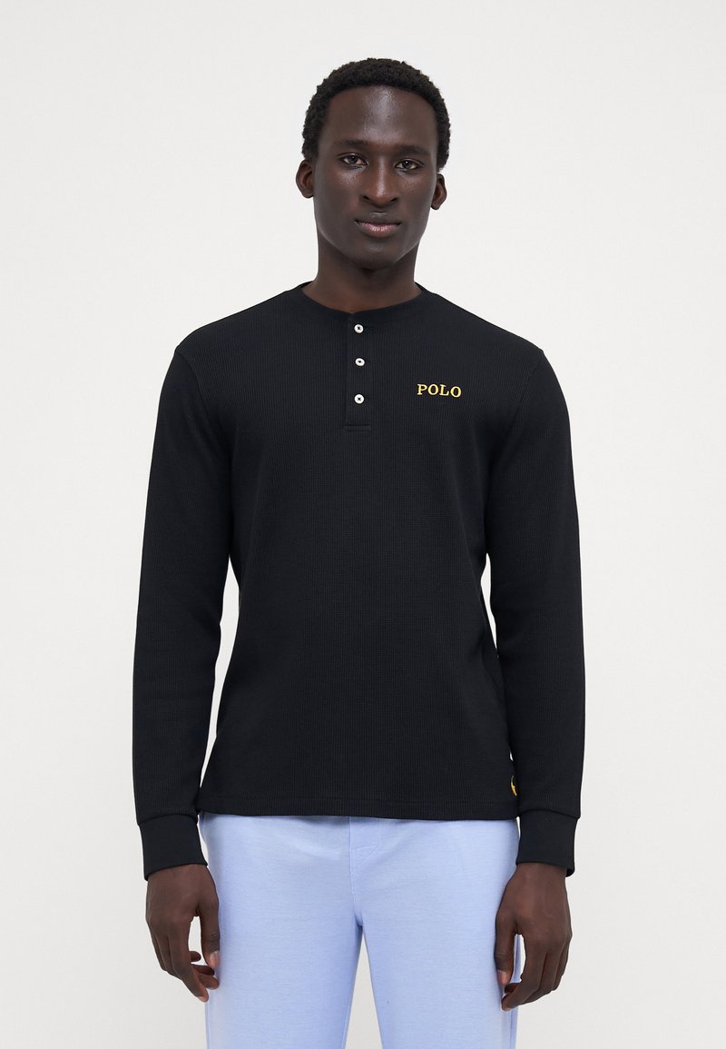 Camisola henley preta de mangas longas com textura canelada, com abertura de três botões e logótipo amarelo "POLO" no lado esquerdo do peito. Combinada com calças azul claro.