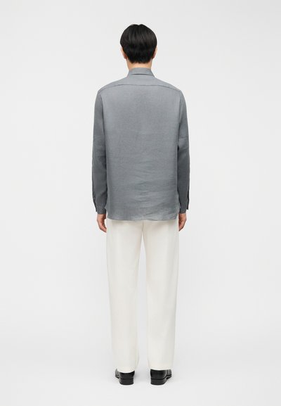 Άνδρας που στέκεται με την πλάτη στραμμένη, φοράει μακρυμάνικο γκρι πουκάμισο, λευκό παντελόνι και μαύρα παπούτσια, σε λευκό απλό φόντο.