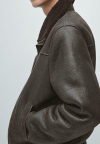 Sideteknisk billede af en person iført mørkebrun læderjakke med tekstur og blød krave, hænderne i lommerne mod en ensartet lys baggrund.