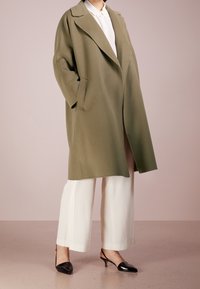 Manteau en laine vert olive avec des revers surdimensionnés, des poches latérales et une ouverture à l'avant. Associé à un pantalon large blanc et des talons vernis noirs.