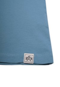 Vaaleansininen puuvillapaita, jossa on pehmeä kangas, jossa on valkoinen etiketti mustalla painatuksella ja logo alahelassa.