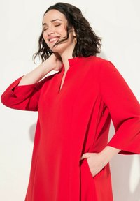 Vestido rojo con un cuello alto, mangas tres cuartos, bolsillos laterales y una textura suave y fluida, situado sobre un fondo neutro.