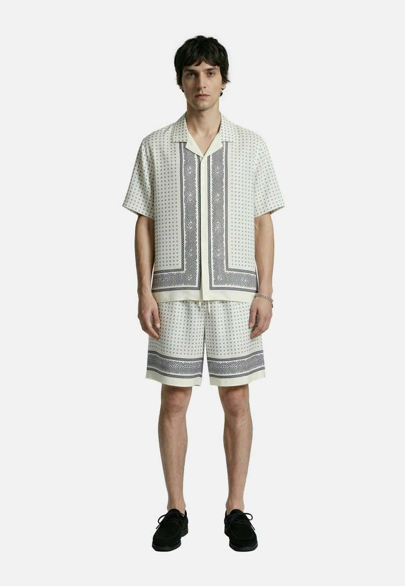 Hombre con camisa blanca de manga corta y pantalones cortos a juego con patrones geométricos negros, de pie con zapatos negros contra un fondo liso.