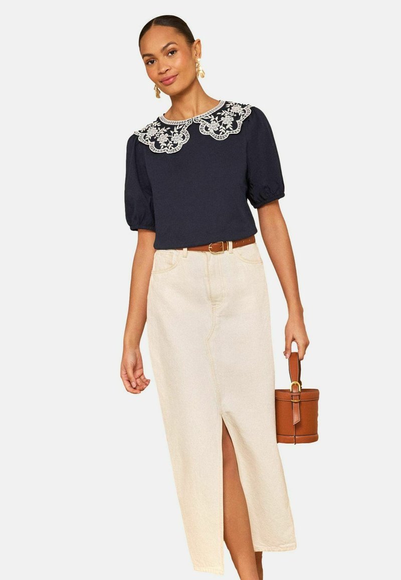 Kobieta w granatowej bluzce z białym koronkowym kołnierzem w kwiatowy wzór, beżowej spódnicy z wysokim stanem i przednim rozcięciem, trzymająca brązową skórzaną torebkę.