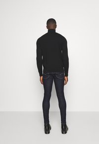 Suéter de cuello alto negro sobre jeans ajustados azul oscuro, con una silueta ceñida y textura suave. La modelo está de espaldas a la cámara.