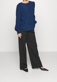 Blusa a maniche lunghe con stampa blu e vestibilità ampia, collo alto; abbinata a pantaloni neri a gamba larga e décolleté neri a punta.