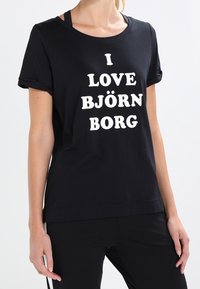 Camiseta de algodón negra con mangas enrolladas y texto blanco que dice "ME ENCANTA BJÖRN BORG". Corte recto, textura suave, diseño minimalista.