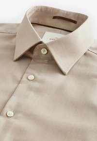Beige Hemd mit Button-Down-Kragen, zwei weißen Knöpfen an der Knopfleiste und einem strukturierten Stoff mit einem feinen Muster.