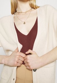 Vrouw in laagjes gouden kettingen, met een bordeauxrode gebreide top, een beige cardigan en tan kleurige broek, die de randen van de cardigan met beide handen vasthoudt.