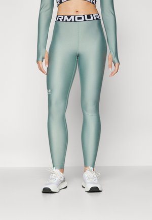Leggings de tiro alto y ajustados en material mate color verde azulado con cinturilla negra que dice "ARMOUR"; presenta el logo de Under Armour cerca del tobillo.