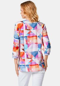Camicia a maniche tre quarti con motivo geometrico colorato. Il materiale sembra leggero e presenta righe ai polsini. Sfondo bianco.