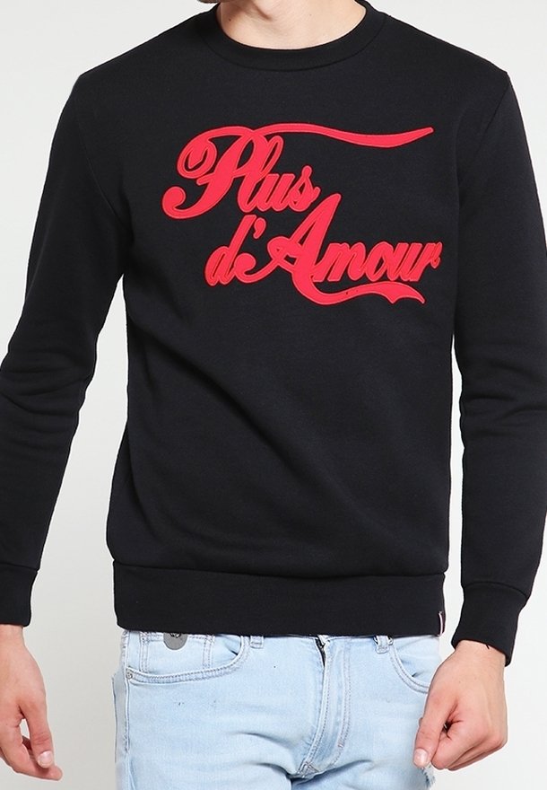 Chemise noire à manches longues avec texte rouge en cursive "Plus d'Amour" portée par une personne portant un jean bleu clair.