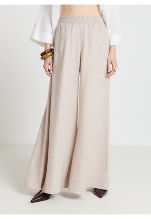 Donna che indossa pantaloni beige a gamba larga con cintura elastica, maniche di camicia bianca visibili, scarpe marroni a punta e braccialetti spessi al polso.