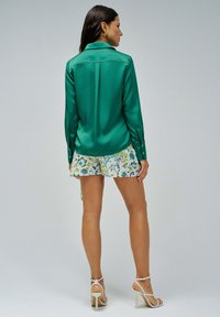 Blusa de satén verde con mangas largas y puños con botones, combinada con pantalones cortos blancos con un patrón floral. Textura suave y corte relajado.