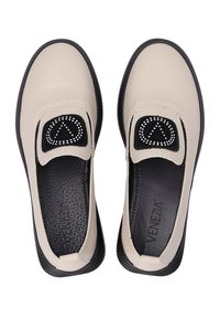 Beige Slipper mit schwarzen Akzenten, Gummisohle, strukturiertem Fußbett und dekorativen dreieckigen Nieten auf einem schwarzen Stoffpatch.