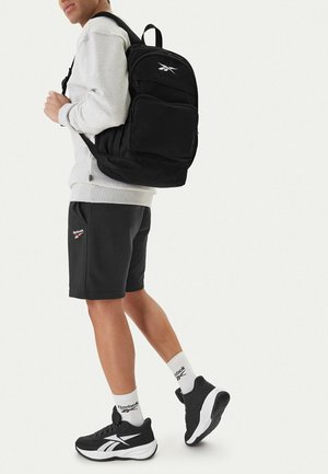 Osoba nosząca czarne szorty Reebok, czarno-białe trampki, białe skarpetki Reebok, szarą bluzę oraz niosąca czarny plecak Reebok.