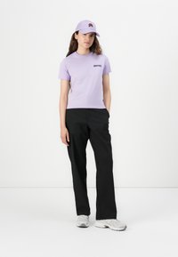 Kratka vijolična majica s kratkimi rokavi in črnim logotipom, združena s črnimi hlačami in belimi supergami. Dopolnjena z vijolično kapo z rožnim vzorcem.