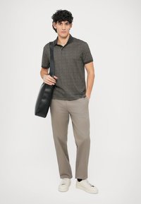 Čierne polo tričko so vzorom, béžové nohavice, biele tenisky. Model drží čiernu kabelku na rameno, pózuje na bielom pozadí.