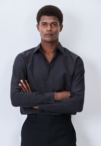 Schwarzes langärmeliges Hemd mit einem dezenten, strukturierten Muster, klassischem Kragen, Knopfverschluss und schwarzen Knöpfen. Kombiniert mit schwarzer Hose.