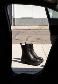 Bottes plateformes épaisses noires debout sur un trottoir ensoleillé vues à travers le cadre d'une porte de voiture.