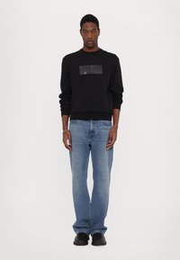 Felpa nera con un patch quadrato lucido sul petto, abbinata a jeans azzurri e scarpe nere. Il modello sta dritto con un'espressione neutra.