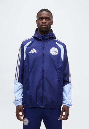 Homme portant une veste et un pantalon de sport bleu marine et bleu clair avec des écussons de logo, debout devant un fond blanc uni.