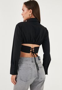 Woman with long brown hair wearing a black cropped shirt with back lace-up detail and gray high-waisted jeans, facing sideways.