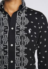 Svart skjorta med knappar, prydd med vita broderade mönster, diamantformer och en liten logotyp på bröstet. Med krage och texturerat tyg.