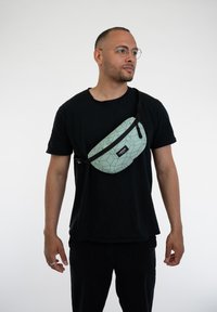 Borsa a spalla verde chiaro con motivo geometrico e dettagli neri, realizzata in materiale liscio, indossata crossbody.