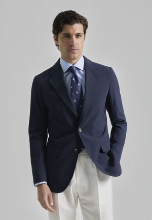 Blazer navy hecho de tejido suave, con solapas muestradas, dos bolsillos frontales y cierre de un solo botón, acompañado de una camisa azul claro y una corbata con estampado.