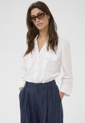 Chemise blanche boutonnée avec deux poches frontales, associée à un pantalon bleu marine à fines rayures. Le modèle porte des lunettes de soleil écaille de tortue. Texture lisse.