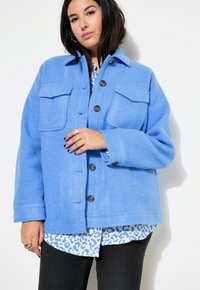 Camisa azul de forro polar con dos bolsillos frontales, cierres de botones y una textura suave, superpuesta sobre una blusa estampada. Jeans negros visibles por debajo.