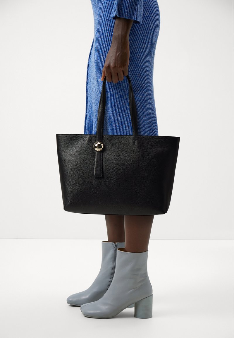 Personne portant une robe bleue et des bottines gris clair tenant un grand sac fourre-tout en cuir noir avec un pendentif rond doré.