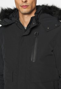 Sort parka jakke med hætte trimmet med pels, lynlåslomme på brystet og to forlommer. Har trykknap- og lynlåslukninger samt en elegant mat finish.